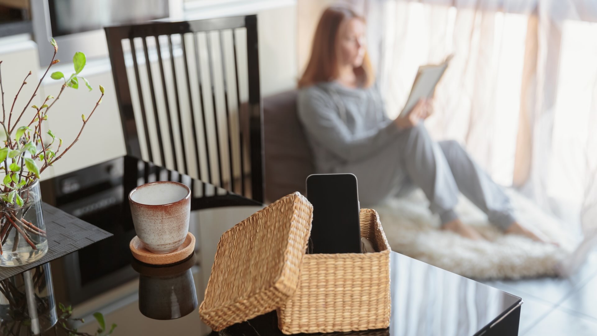 Ein Smartphone liegt in einem Korb verwahrt auf einem Glastisch. Im Hintergrund sitzt eine Person auf dem Boden am Fenster und liest ein Buch.