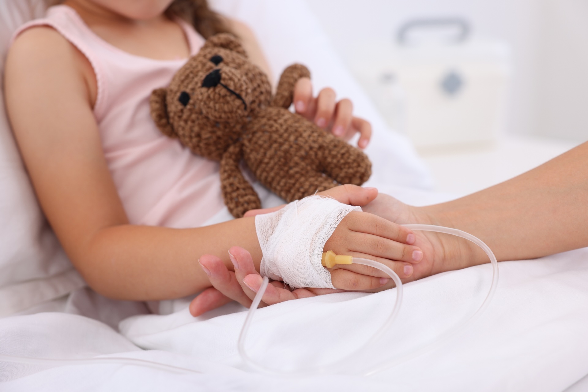 Ein M&auml;dchen liegt in einem Krankenhausbett und h&auml;lt einen Teddy in der Hand. Eine erwachsene Person h&auml;lt die Hand des M&auml;dchens, in der sich ein medizinischer Zugang befindet.