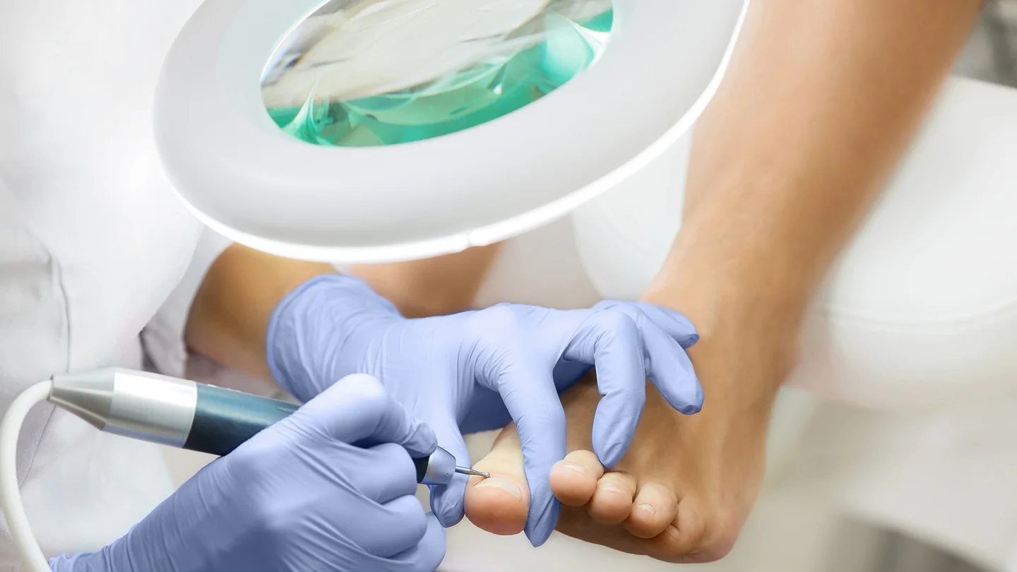 Eine Podologin behandelt mit einem Fußpflegegerät einen Fuß unter einer Lupenlampe.