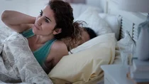 Eine Frau richtet sich in einem Bett auf und fasst sich an die Schulter. 