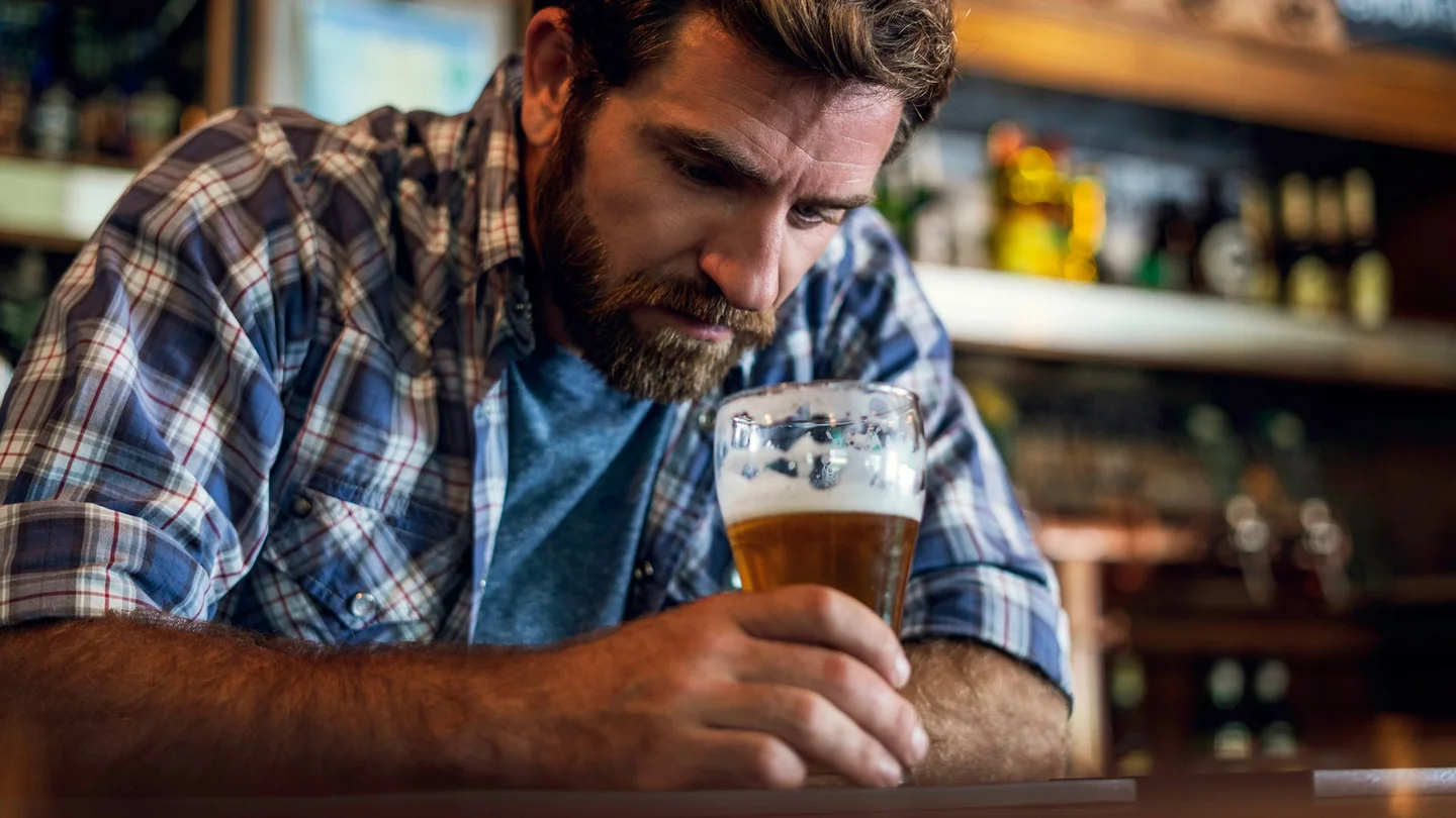 Ein Mann sitzt an einer Bar, h&auml;lt ein Bierglas in der Hand und schaut in den Glas.