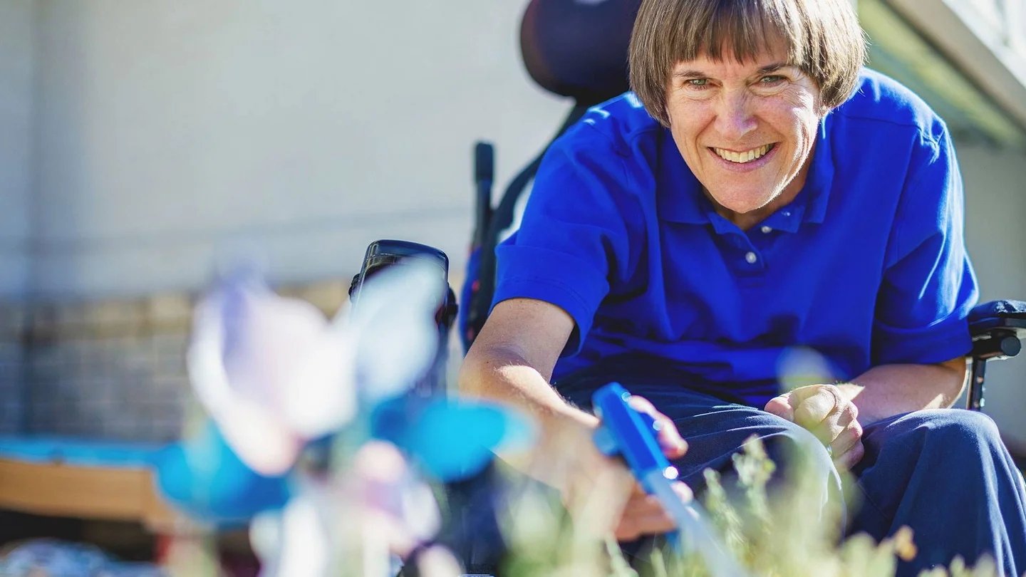 Eine Frau mittleren Alters sitzt im Rollstuhl und pflegt ihre Blumen mit einem Handgreifer.