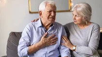 Ein &auml;lterer Herr sitzt auf dem Sofa und hat seine Hand mit schmerzverzerrtem Gesicht auf seine Brust gelegt. Eine &auml;ltere Dame sitzt neben ihm, legt ihm f&uuml;rsorglich ihre H&auml;nde auf Schulter und Arm und sieht ihn besorgt an.