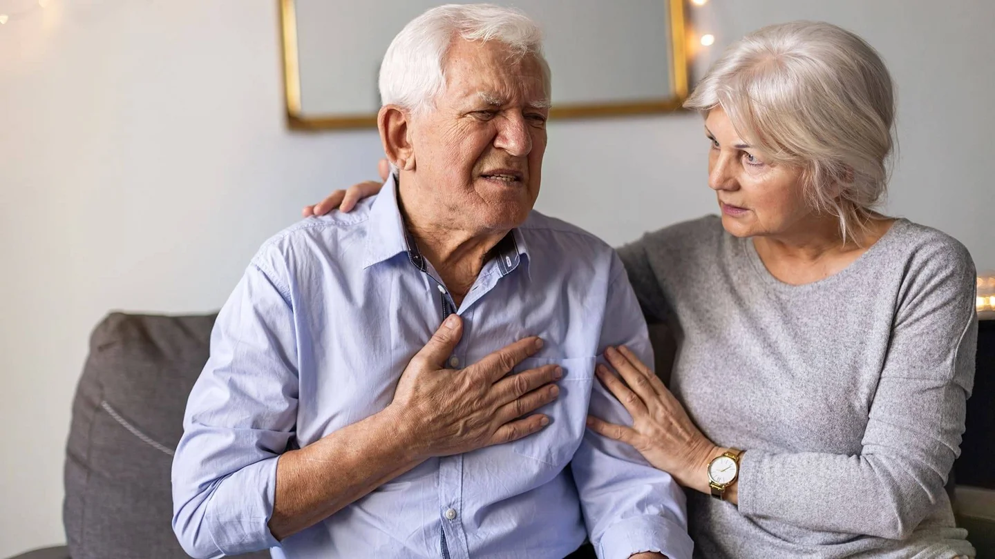 Ein &auml;lterer Herr sitzt auf dem Sofa und hat seine Hand mit schmerzverzerrtem Gesicht auf seine Brust gelegt. Eine &auml;ltere Dame sitzt neben ihm, legt ihm f&uuml;rsorglich ihre H&auml;nde auf Schulter und Arm und sieht ihn besorgt an.