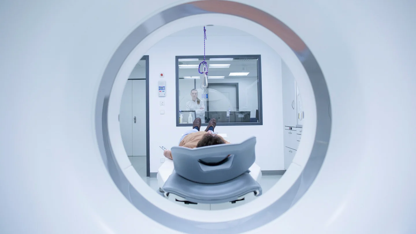 CUP-Syndrom (Krebs bei unbekanntem Prim&auml;rtumor): Ein Patient liegt vor einer MRT-R&ouml;hre. Im Hintergrund steht eine &Auml;rztin hinter einer Glasscheibe und beobachtet den Patienten.