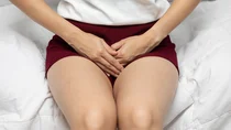 Harnwegsinfektion Zystitis: Eine junge Frau sitzt auf einem Bett und fasst sich mit beiden H&auml;nden an den Schritt.
