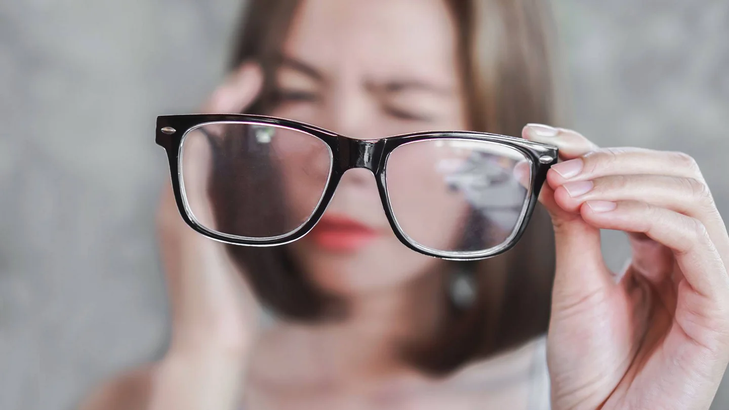 Verschwommenes Gesicht einer Frau, die sich benommen an den Kopf fasst. Sie h&auml;lt eine Brille in den Vordergrund, die scharf zu sehen ist.