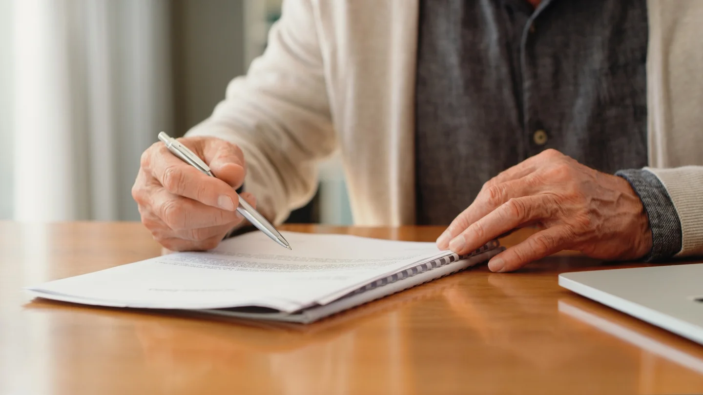 Eine ältere Person sitzt am Tisch und unterschreibt ein Dokument.