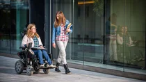 Zwei junge Frauen mit Büchern bewegen sich vorwärts, eine von Ihnen sitzt in einem Rollstuhl. 