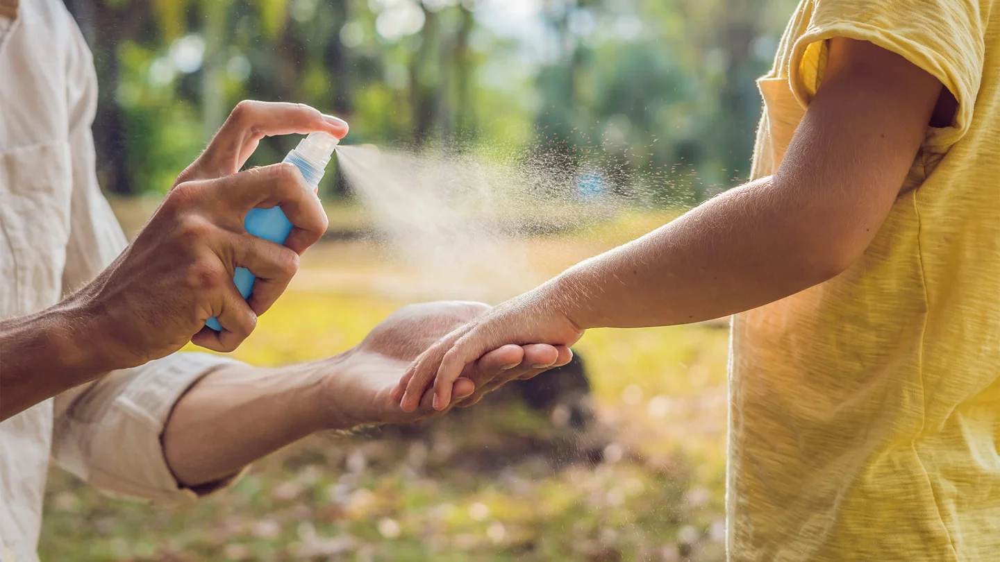 Exotische Infektionskrankheiten: Nahaufnahme von Händen und Armen zweier T-Shirt tragender Personen vor einem parkähnlichem Hintergrund. Eine Person hält den Arm der anderen und sprüht Mückenschutz darauf.
