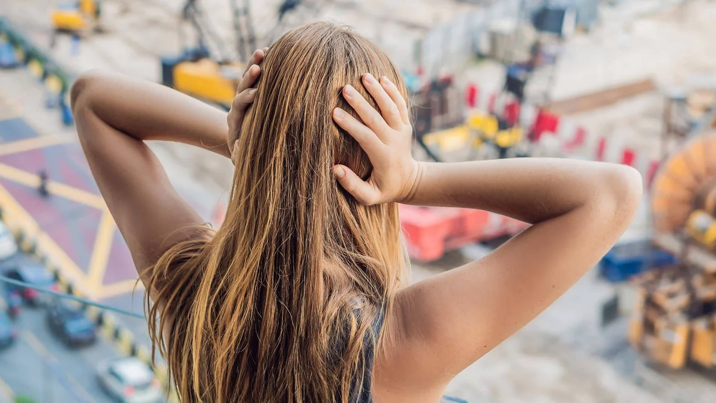 Von hinten ist eine junge weibliche Person zu sehen, die sich die Ohren zuhält. Vor ihr verschwommen eine städtische Umgebung mit Baustellen und Autoverkehr.
