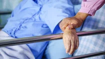 Eine Person liegt in einem Bett mit einem Bettgitter aus Metall. Sie hat den Arm auf das Bettgitter gelegt. Eine weitere Person berührt diesen Arm.