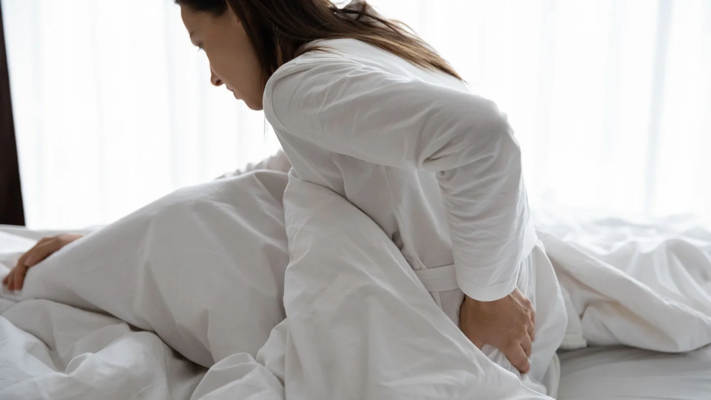 Frau sitzt im Bett und fasst sich an den unteren R&uuml;cken.