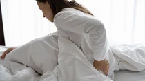 Frau sitzt im Bett und fasst sich an den unteren R&uuml;cken.