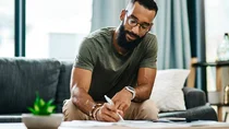 Ein Mann sitzt am Wohnzimmertisch. Er hält einen Stift in der Hand und füllt ein Formular aus.