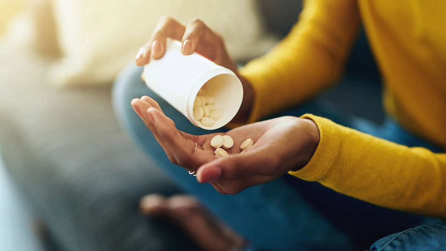 Nahaufname: Eine junge Frau sch&uuml;ttet mehrere Tabletten aus einer gr&ouml;&szlig;eren Medikamentendose in ihre offene Hand.