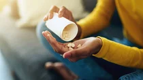 Nahaufname: Eine junge Frau sch&uuml;ttet mehrere Tabletten aus einer gr&ouml;&szlig;eren Medikamentendose in ihre offene Hand.