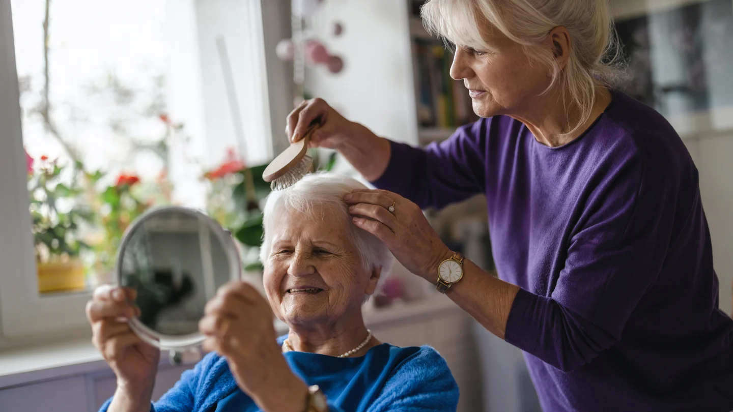 Eine Frau k&auml;mmt ihrer &auml;lteren Mutter die Haare. Die Mutter betrachtet sich dabei l&auml;chelnd im Spiegel.