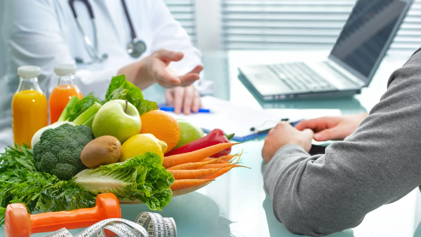 Eine Ernährungsberaterin und ein Patient führen ein Gespräch neben einem gefüllten Obstkorb.