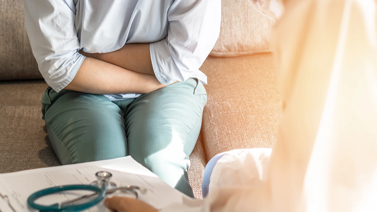 Gonorrhö: Eine Frau auf einem Sofa. Sie hält sich mit beiden Händen den Bauch. Sie wirkt angespannt. Ihr gegenüber sitzt ein Arzt.