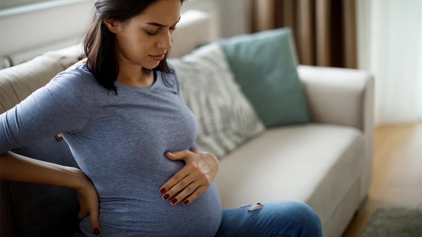 Typische Schwangerschaftsbeschwerden: Eine hochschwangere junge Frau sitzt mit geschlossenen Augen auf dem Sofa. Sie h&auml;lt sich den Bauch und den R&uuml;cken. Sie scheint R&uuml;ckenschmerzen zu haben.