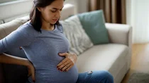 Typische Schwangerschaftsbeschwerden: Eine hochschwangere junge Frau sitzt mit geschlossenen Augen auf dem Sofa. Sie h&auml;lt sich den Bauch und den R&uuml;cken. Sie scheint R&uuml;ckenschmerzen zu haben.
