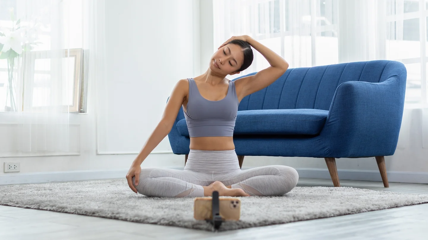 Eine Frau sitzt auf dem Boden vor ihrem Smartphone und macht Entspannungs- und Dehnungsübungen. 