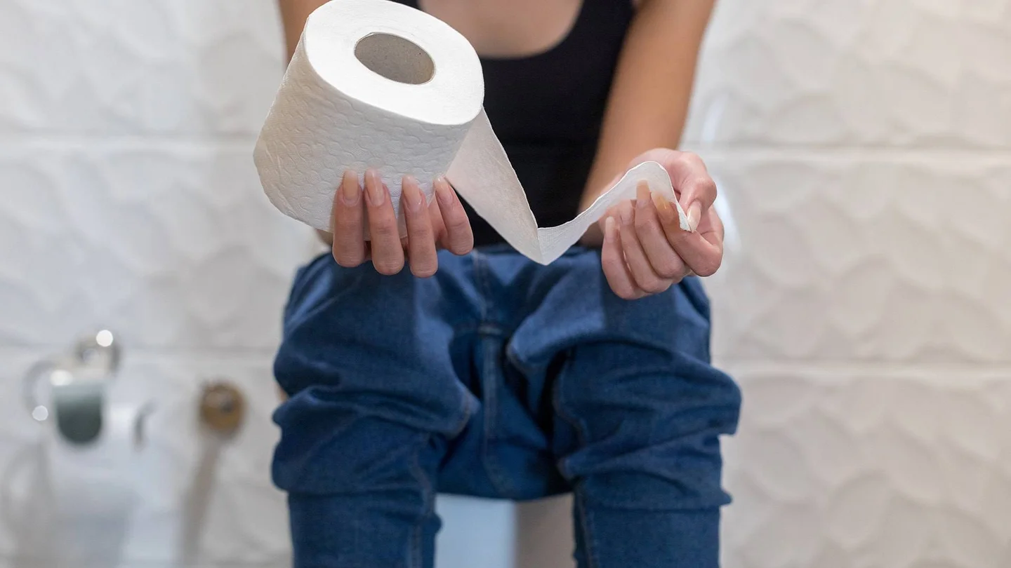 Eine junge Frau sitzt vorgebeugt auf der Toilette und h&auml;lt eine Rolle Toilettenpapier in der Hand.