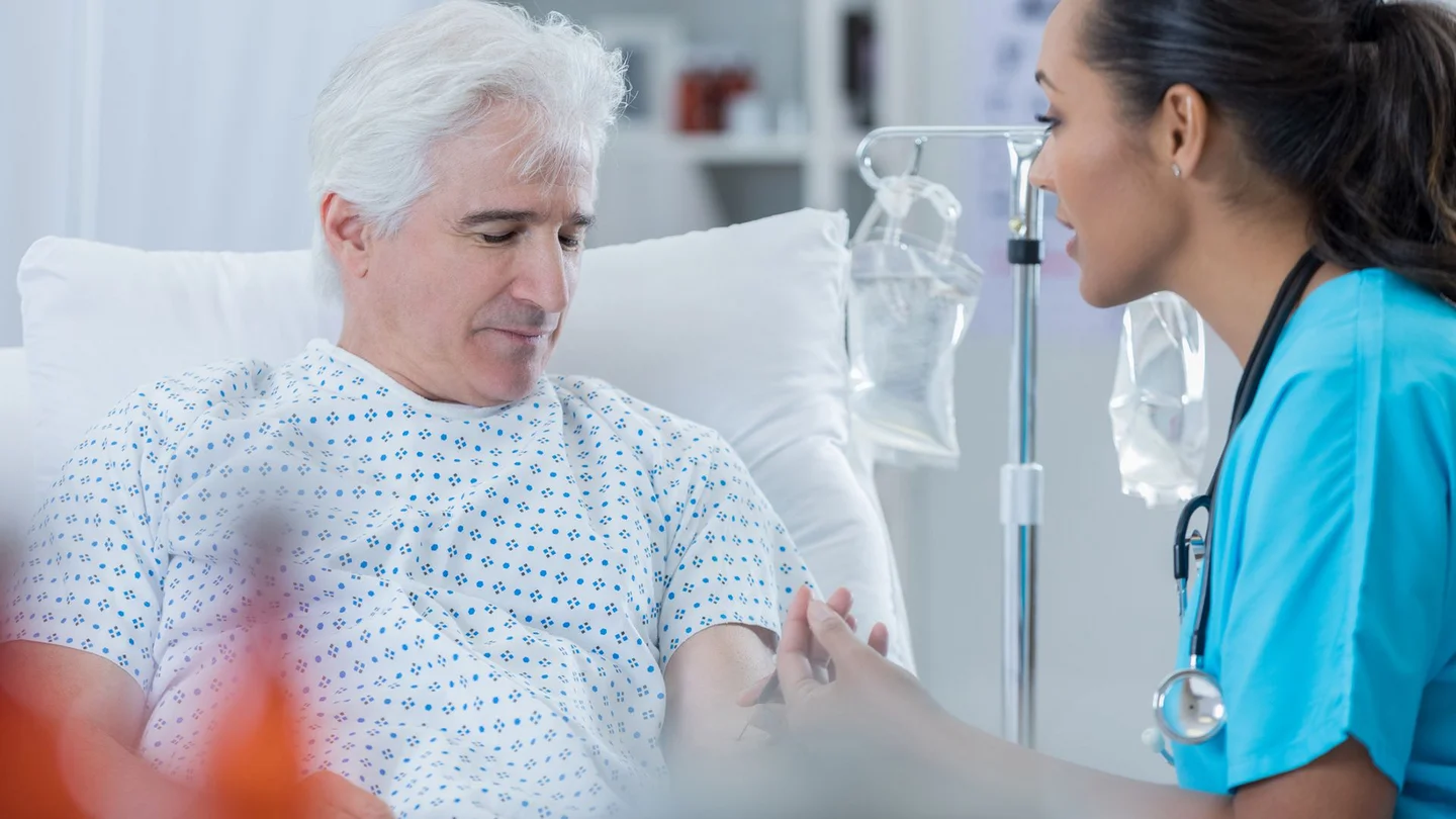 Prostatakrebs: Ein &auml;lterer Mann sitzt in einem Krankenhausbett. Er schaut nach unten, ihm gegen&uuml;ber sitzt eine Krankenschwester. Sie schaut ihn an und entnimmt ihm Blut.
