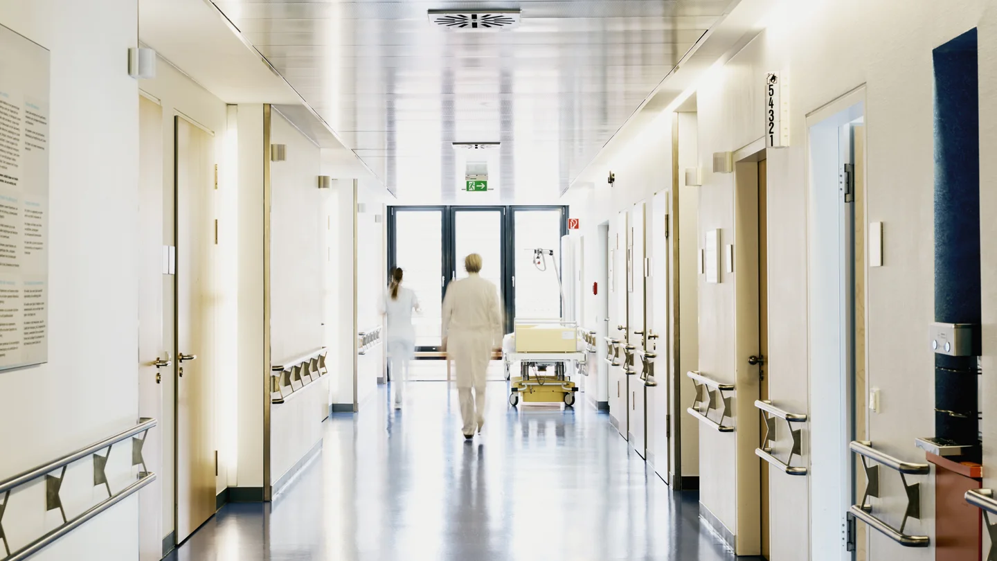 Krankenhauspersonal in weißer Arbeitskleidung läuft durch einen hellen Krankenhausflur.