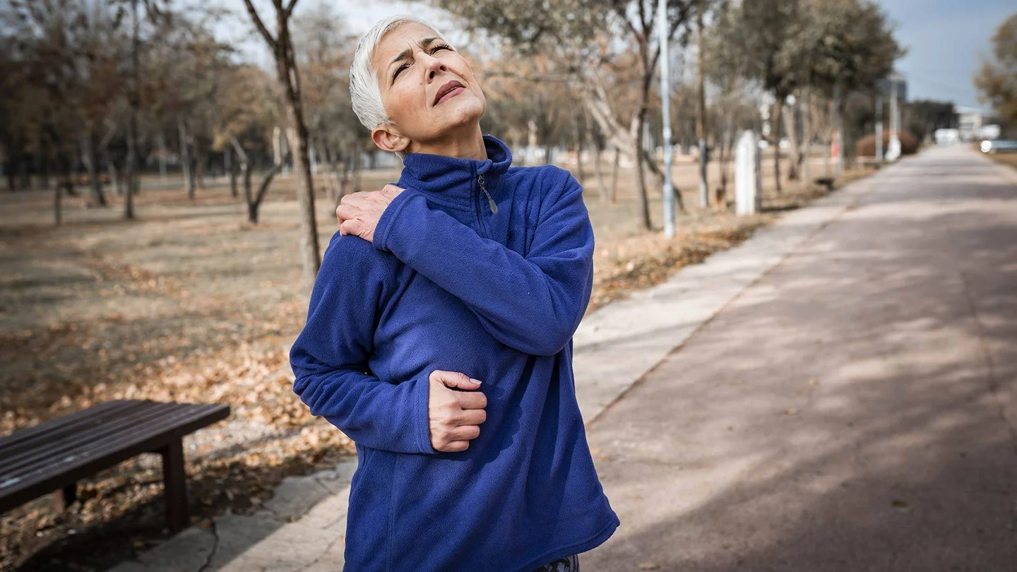 Eine Frau mit grauen Haaren beim Joggen: Sie greift sich mit der linken Hand an die rechte Schulter, die offenbar stark schmerzt.