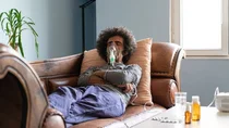 Ein Mann liegt auf dem Sofa. Er ist über eine Maske an ein Beatmungsgerät angeschlossen. Auf dem Tisch neben ihm stehen Medikamente.