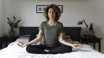 Gesund mit Entspannung: Eine Frau sitzt auf ihrem Bett im Schneidersitz und meditiert. Sie hat beide Hände auf ihren Knien abgelegt und die Hände in Chinmudra-Haltung. Sie hat die Augen geschlossen und atmet aus.