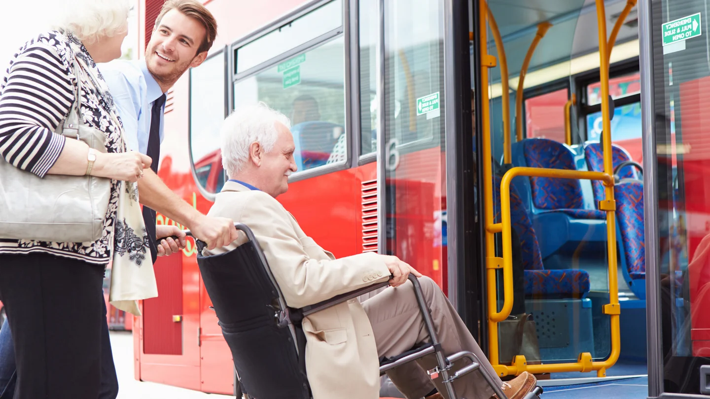 Ein junger Busfahrer schiebt einen &auml;lteren Herrn im Rollstuhl &uuml;ber eine Rampe in den Bus.