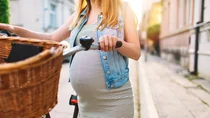 Eine junge hochschwangere Frau schiebt ihr Fahrrad über die Straße. Sie ist sommerlich gekleidet und die Sonne scheint.