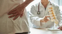 Frau greift sich an den unteren Rücken, ihr gegenüber sitzt ein Arzt mit Wirbelsäulenmodell.