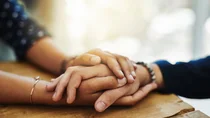 Eine Person hält über den Tisch hinweg tröstend die Hand einer anderen Person in ihren beiden Händen.