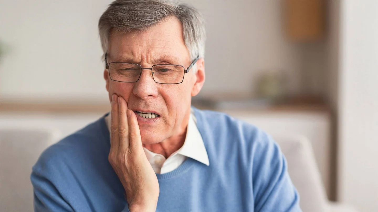 Ein gepflegter älterer Herr fasst sich besorgt an die rechte Wange; sein Gesicht ist schmerzverzerrt.