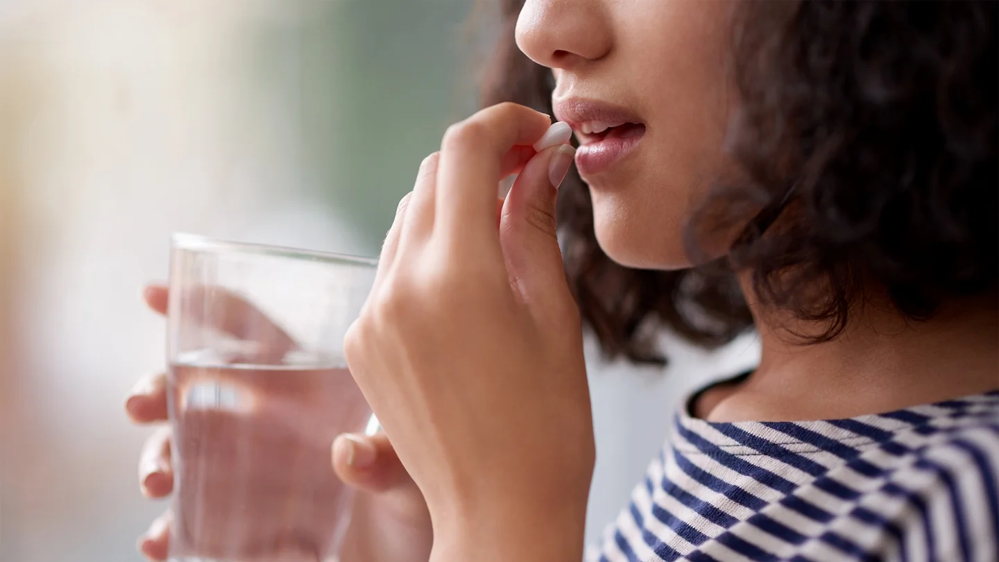 Notfallverh&uuml;tung: Eine junge Frau h&auml;lt ein Wasserglas in der einen Hand. Mit der anderen Hand f&uuml;hrt sie eine Tablette zum halb ge&ouml;ffneten Mund.