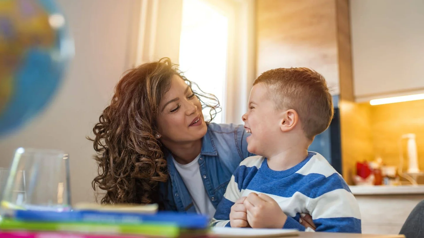 Ein kleiner Junge sitzt am Küchentisch und dreht sich lachend zu einer Frau um, die sich von der Seite zu ihm gebeugt hat und ihn freundlich anblickt.