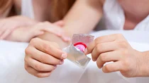 STI: Ein Mann &ouml;ffnet eine silberne Kondomverpackung. In der Verpackung steckt ein rotes Kondom.