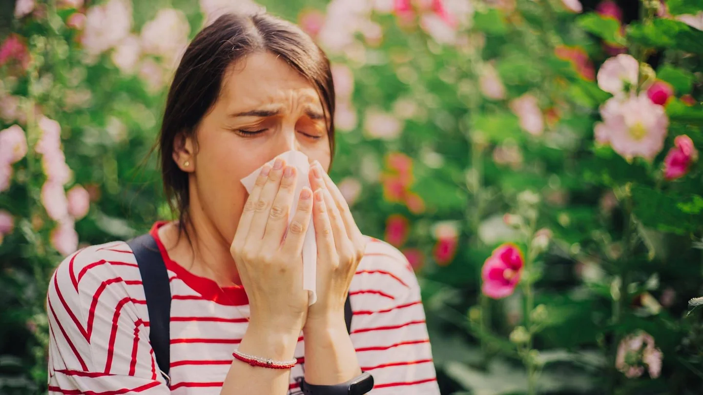 Eine Frau im T-Shirt schnäuzt sich in ihr Taschentuch. Im Hintergrund sind blühende Blumen zu sehen.
