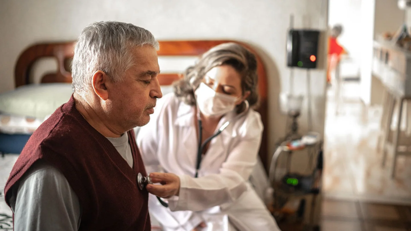 Ein &auml;lterer Mann sitzt auf einem Krankenbett. Sein Gesichtsausdruck deutet auf k&ouml;rperliche Schw&auml;che hin. Eine &Auml;rztin mit Mundschutz h&ouml;rt sein Herz mit dem Stethoskop ab.