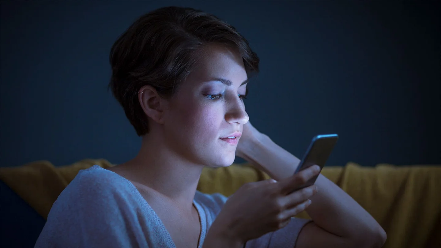 Eine junge Frau sitzt im Halbdunkeln auf dem Sofa. Sie h&auml;lt ein Mobiltelefon nahe am Gesicht, und schaut konzentriert auf den Bildschirm. Ihr Gesicht wird vom bl&auml;ulichen Licht des Displays angestrahlt.
