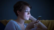 Eine junge Frau sitzt im Halbdunkeln auf dem Sofa. Sie h&auml;lt ein Mobiltelefon nahe am Gesicht, und schaut konzentriert auf den Bildschirm. Ihr Gesicht wird vom bl&auml;ulichen Licht des Displays angestrahlt.