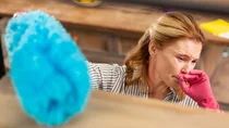 Hausstauballergie: Eine Frau wischt Staub und hält sich die Hand ins Gesicht. Ihre Augen sind gerötet.