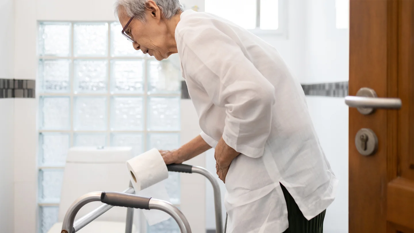 Colitis ulcerosa: Eine ältere Dame ist auf dem Weg zur Toilette. Sie stützt sich auf ihren Rollator und hält sich den Bauch.