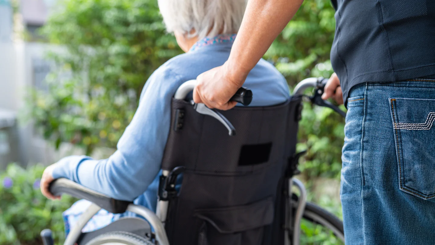Eine Seniorin sitzt in einem Rollstuhl, der von einem Mann geschoben wird.