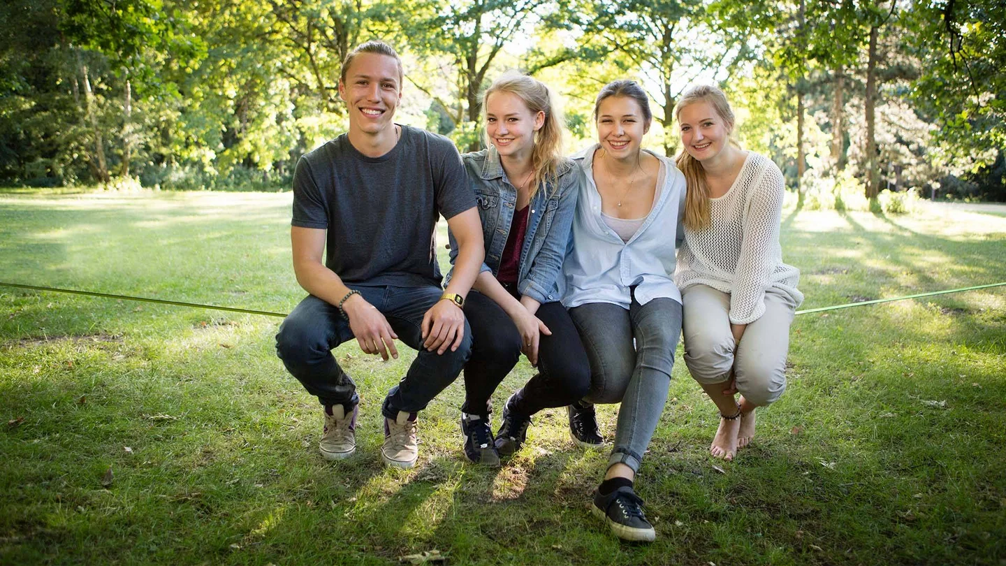 Vier junge Menschen sitzen im Park auf einem gespannten Seil.