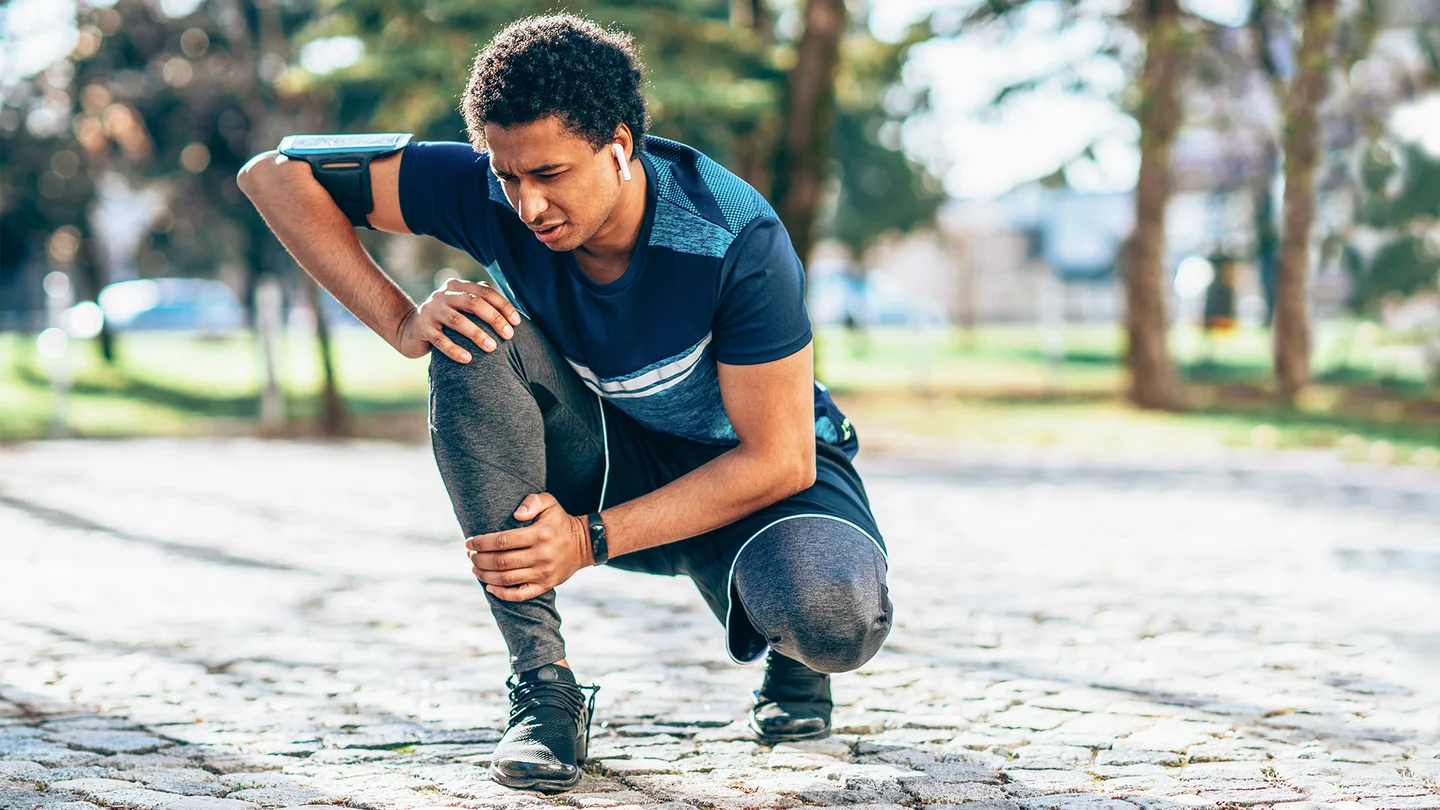 Vorderer Knieschmerz: Ein junger Mann in Sportkleidung kniet im Freien. Er hält sich das Knie und verzieht das Gesicht. Er scheint sein Lauftraining aufgrund von Schmerzen unterbrochen zu haben.